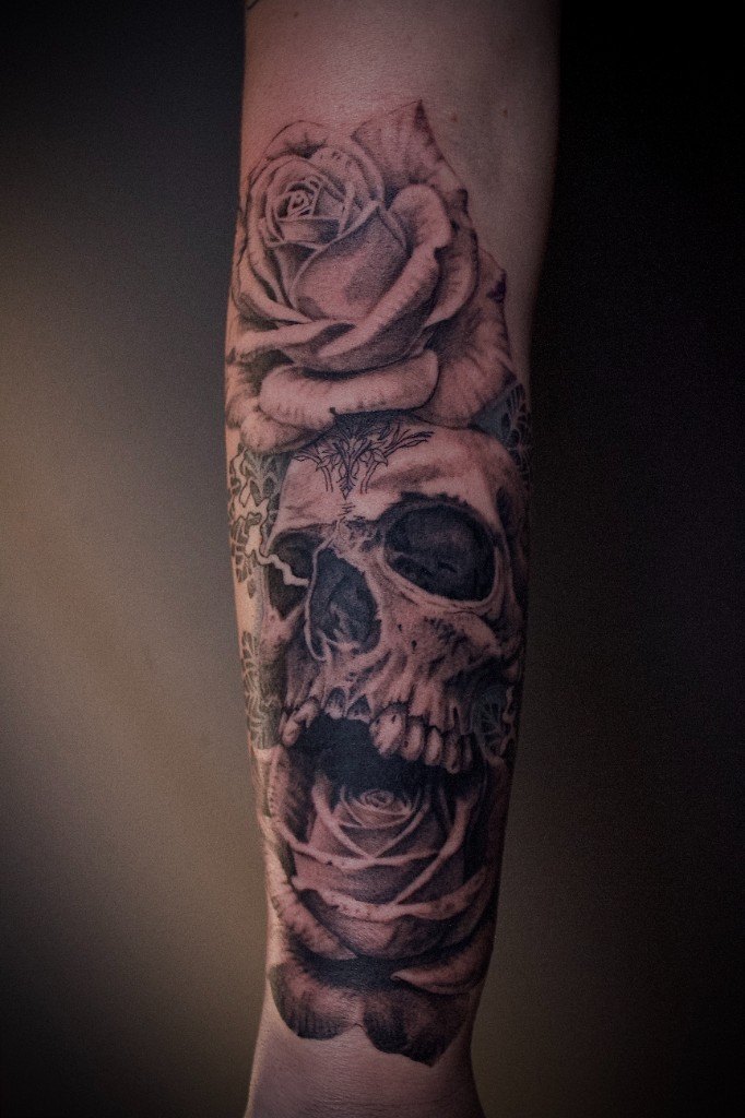 Black and grey realism tattoo of a screaming skull set between two roses on a forearm