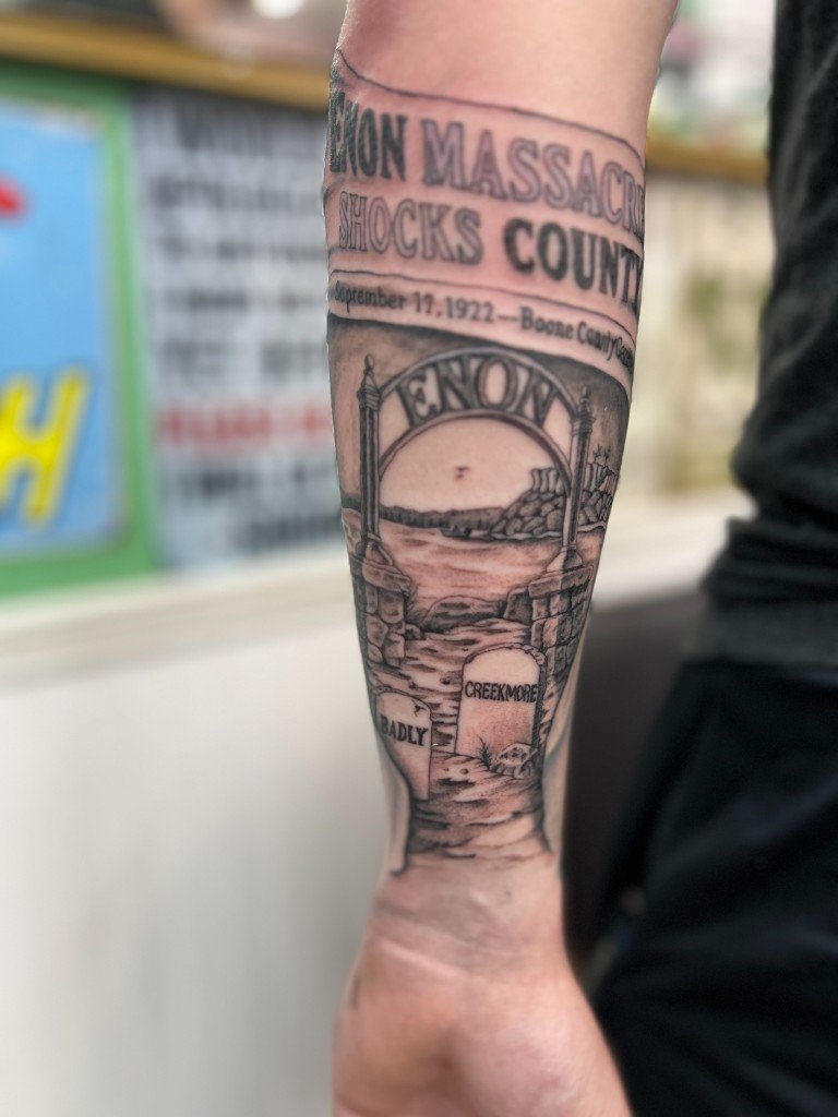 Black and grey tattoo of a 1922 newspaper headline above cemetery gates and headstones, inner forearm