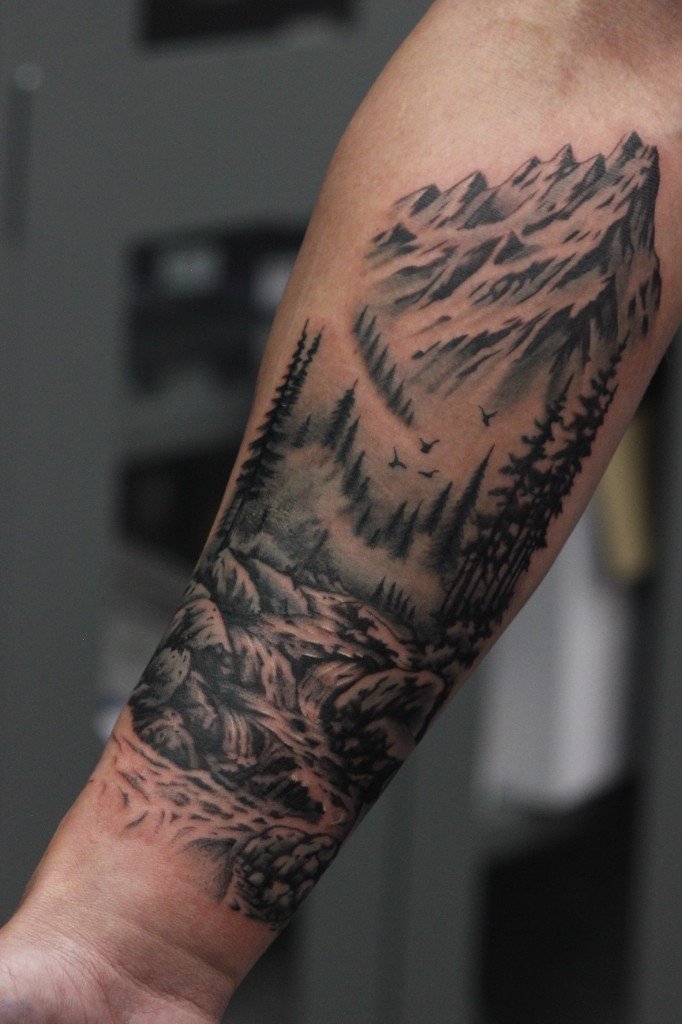 Black and grey mountain range and pine forest landscape tattoo wrapping a forearm